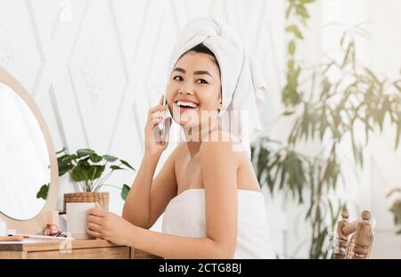 Happy asian lady talking on phone, sitting next to mirror Stock Photo