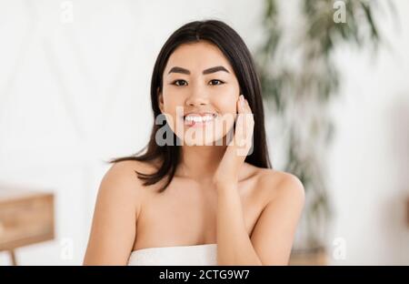 Cheerful asian woman wrapped in towel touching her face Stock Photo