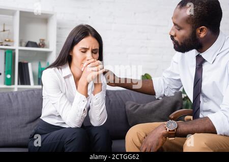 african american psychologist calming upset woman sitting on sofa with closed eyes and clenched hands Stock Photo