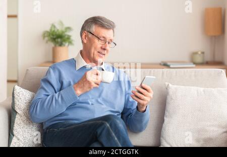 Senior Man Using Smartphone Having Coffee Sitting On Couch Indoor Stock Photo