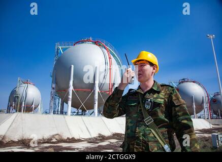 Oil refinery and gas processing plant. Asian Security guard with radio set on liquefied gas (LPG) storage sphere background. Stock Photo
