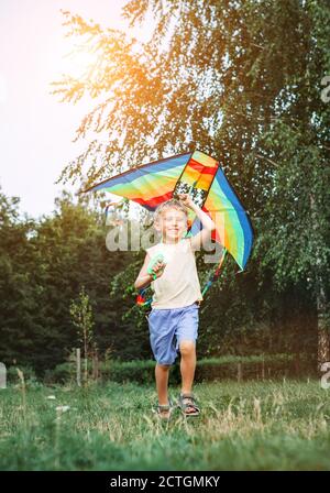 Cheerful little boy running with a multicolored kite on the city park green grass meadow. Funny childhood concept image. Stock Photo