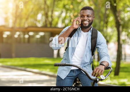 Smiling Black Guy Talking On Cellphone While Riding Bicycle Outdoors Stock Photo