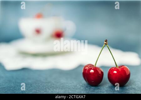 Two red, delicious cherries in front of an old porcelain cup on the table. Blue background Stock Photo