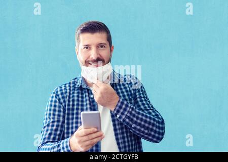 Confident mature man with big smile and open protective face mask using smartphone against wall Stock Photo