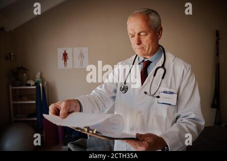 Senior male doctor holding clipboard checking reports in modern home hospital Stock Photo