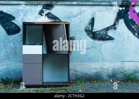An old broken cupboard stands in front of a silver-sprayed wall, bulky waste on the street, broken cupboard on the street Stock Photo