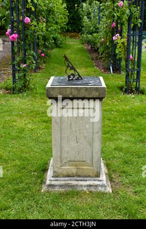 close up of sun dial mounted on a stone pillar set in a country garden with arches of roses behind Stock Photo