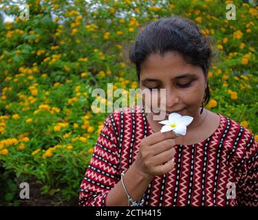 Pretty brunette smells a white flower wearing a designer top and bracelets, with yellow flowers in background during springtime Stock Photo