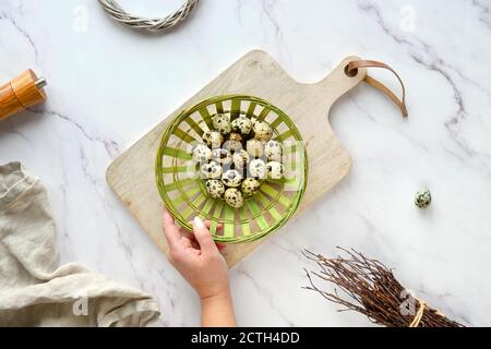 Hand holds rattan plate, basket with quail eggs. Flat lay on white marble table. Stock Photo