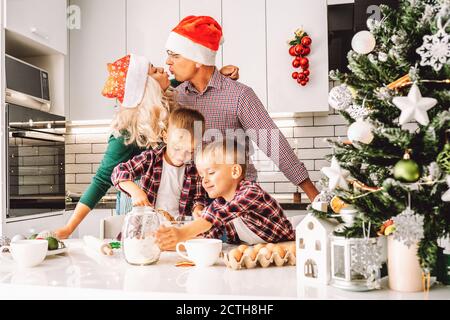 Family of two twins boys and age parents preparing cookies for Christmas eve in light kitchen wearing Santa hats. Stock Photo