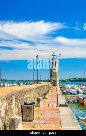 Stone pier mole with lighthouse, street lights and yachts on boat parking port marina in Desenzano del Garda town, blue sky white clouds background, vertical view, Lombardy, Northern Italy Stock Photo