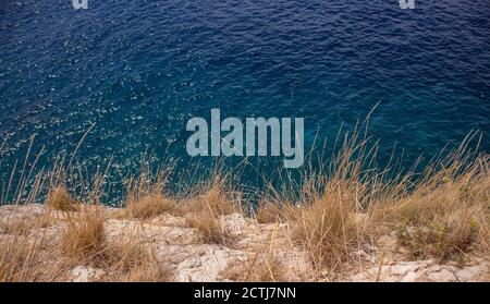 Steep rocky cliff vertically above deep, blue sea off the shore of Hvar island, Croatia, overgrown in dry grass waving in the wind Stock Photo