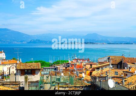 Aerial panoramic view of Desenzano del Garda town with red tiled roof buildings, Garda Lake water surface, Monte Baldo mountain range, Sirmione peninsula, blue sky background, Lombardy, Northern Italy Stock Photo