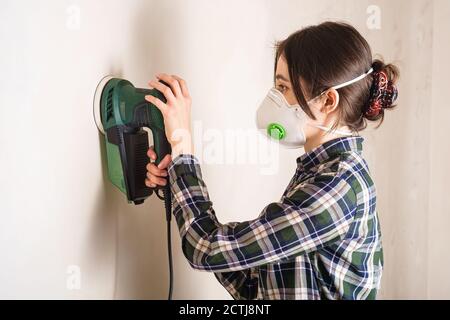 Woman in protective mask working with electric sander to smooth plaster wall surface, room renovation concept Stock Photo