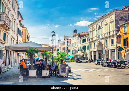 Chioggia, Italy, September 16, 2019: people in street restaurant , riding cars, colorful multicolored buildings in historical town centre, blue sky background in summer day, Veneto Region Stock Photo