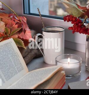 An iron cup of coffee with a spoon on the windowsill among a vase with branches and viburnum berries, a sweater, an open book and a candle. Photo for Stock Photo