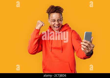 Freestyle. Mulatto freckled guy in hoodie standing isolated on yellow watching video on smartphone shouting excited Stock Photo