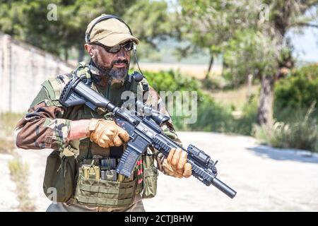 Private military company mercenary, brutal looking special forces fighter in battle uniform and plate carrier, wearing radio headset and sunglasses, holding service rifle in hands, ready to fight Stock Photo