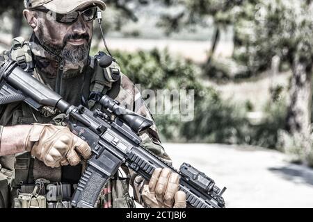 Private military company mercenary, brutal looking special forces fighter in battle uniform and plate carrier, wearing radio headset and sunglasses, holding service rifle in hands, ready to fight Stock Photo