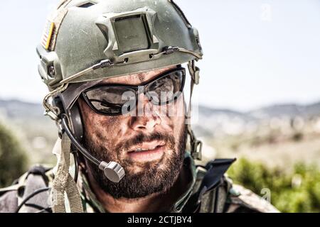 Shoulder portrait of army soldier, special forces fighter, modern warfare combatant with dirty, unshaven face, wearing sunglasses, combat helmet and talking in tactical radio headset during mission Stock Photo