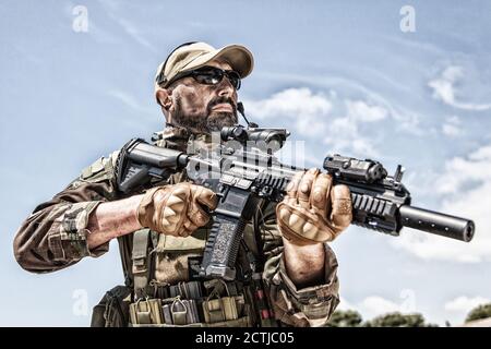 Private military company mercenary, brutal looking special forces fighter in battle uniform and plate carrier, wearing radio headset and sunglasses, holding service rifle in hands, ready to fight Stock Photo