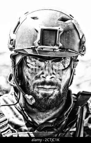 Shoulder portrait of army soldier, special forces fighter, modern warfare combatant with dirty, unshaven face, wearing sunglasses, combat helmet and talking in tactical radio headset during mission Stock Photo