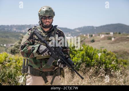 Private military company mercenary, brutal looking special forces fighter in battle uniform and plate carrier, wearing radio headset and sunglasses, holding service rifle in hands, ready to fight Stock Photo