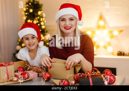 Cheerful pretty mother and her adorable little daughter enjoying beauty of handmade Christmas decoration while sitting at desk, interior of cozy Stock Photo