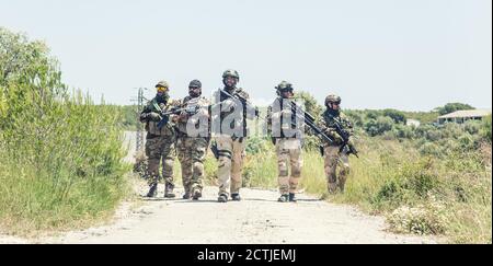 Army soldiers team in combat uniform, load carriers and helmets running on countryside road, aiming guns and sneaking in bush. Special forces infantrymen group moving with cautious at abandoned area Stock Photo
