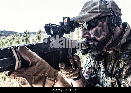 Army special forces soldier, modern combatant with dirty, unshaven face, in combat uniform, glasses and cap, equipped radio headset, aiming service rifle or light machine gun with optical sight Stock Photo