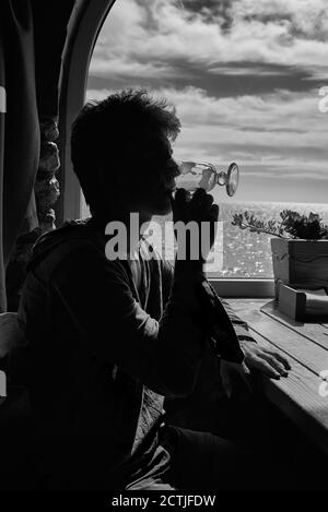 Silhouette of a young man finishing a cappuccino against the background of a window with a view of the sea. Stock Photo