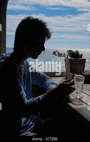 Silhouette of a young man finishing a cappuccino against the background of a window with a sea view. Stock Photo