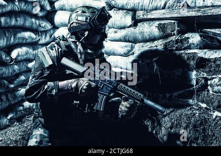 Navy SEALs fighter, army soldier in combat uniform, goggles and battle helmet at night watch, sitting with assault rifle in trench with sandbags, looking in loophole, waiting for enemy attack Stock Photo