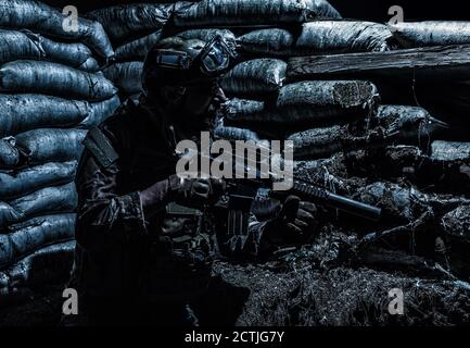 Navy SEALs fighter, army soldier in combat uniform, goggles and battle helmet at night watch, sitting with assault rifle in trench with sandbags, looking in loophole, waiting for enemy attack Stock Photo