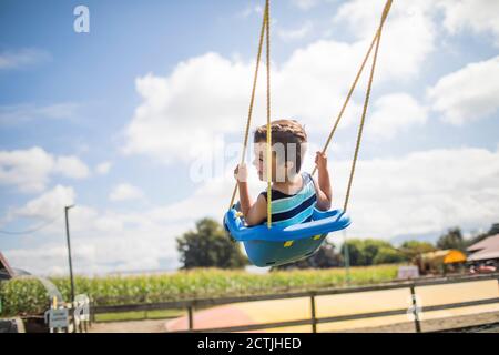 Low angle view of boy swinging outdoors on blue swing at playground. Stock Photo