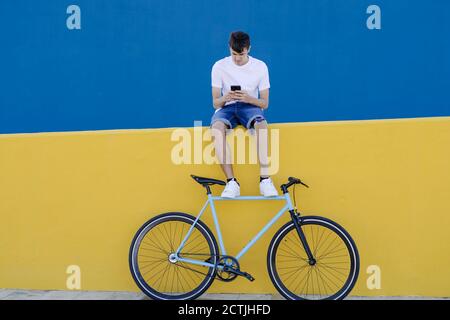 Disabled young male bicyclist sitting on fence and chatting on smartphone Stock Photo