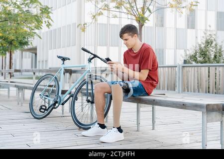 Teen male with leg prosthesis sitting on bench near bike and browsing smartphone Stock Photo