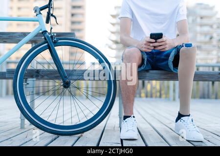 Male cyclist with leg prosthesis sitting on bench and using mobile phone Stock Photo
