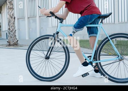 Young male with leg prosthesis sitting on bike and messaging on smartphone in city Stock Photo