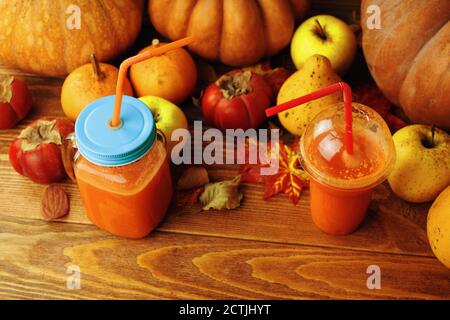 Pumpkin and carrot juice in cups with tubes. Zero waste on the example of glass and plastic cups. Autumn harvest festival. Pumpkins, persimmons, apples and leaves on a wooden background. Stock Photo