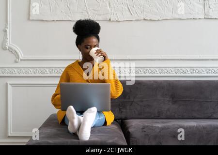 Afro-American biracial woman with afro hairstyle in yellow cardigan sitting on couch, working online on laptop, watching webinar or videos in social m Stock Photo