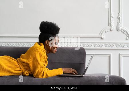 Close up of smiling Afro-American biracial woman with afro hairstyle in yellow cardigan lying on couch, resting, looking at camera webcam and talking Stock Photo