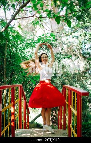 Smiling woman with arms raised practicing ballet on footbridge against trees in park Stock Photo