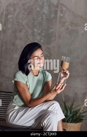 Businesswoman holding hourglass while sitting on chair against wall in office Stock Photo