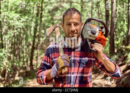 Smiling lumberjack standing with axe and chainsaw in forest Stock Photo