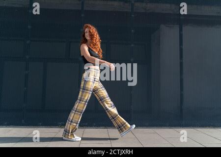 Woman looking over shoulder while walking on sidewalk by fence Stock Photo