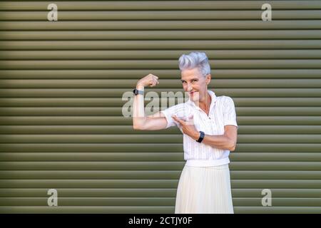 Active senior woman pointing at bicep while flexing muscles while standing against shutter Stock Photo