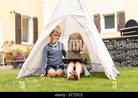 Cute siblings with digital tablet sitting in tent on grass at back yard against house Stock Photo