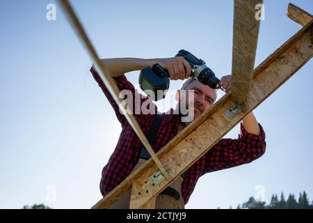 Carpenter drilling wooden plank with power tool against clear sky Stock Photo